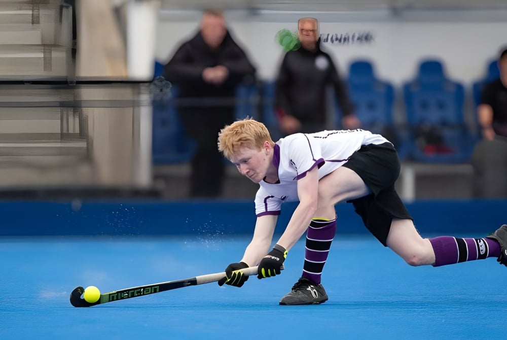 Eine Person spielt Feldhockey auf dem blauen Kunstrasenplatz der Kimbolton School vor einer Gruppe von Zuschauern.