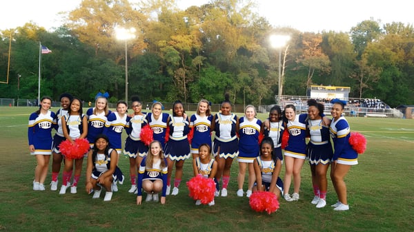 Eine Gruppe Cheerleader in bunten Uniformen posiert gemeinsam auf dem Sportfeld der King George Secondary School.