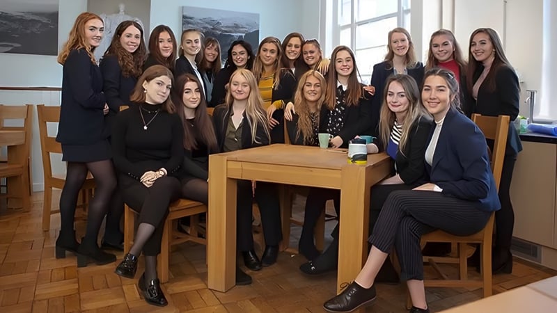 Eine Gruppe junger Frauen von King William's College sitzt formell gekleidet um einen Tisch in einem Klassenraum mit Landschaftsausblick.