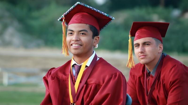 Zwei Absolventen der Kings Christian School tragen rote Talare und Hüte und stehen im Freien auf einer Wiese mit Bäumen im Hintergrund.