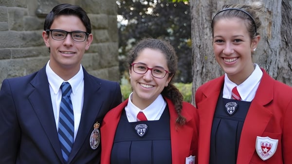 Eine Gruppe von drei Schülerinnen und Schülern steht lachend auf dem Campus der King’s-Edgehill School vor einer Backsteinwand mit Bäumen.