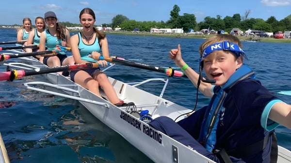 Eine Gruppe von Schülerinnen und Schülern der King's Ely sitzt in einem kleinen Boot auf einem von Bäumen umgebenen Gewässer.