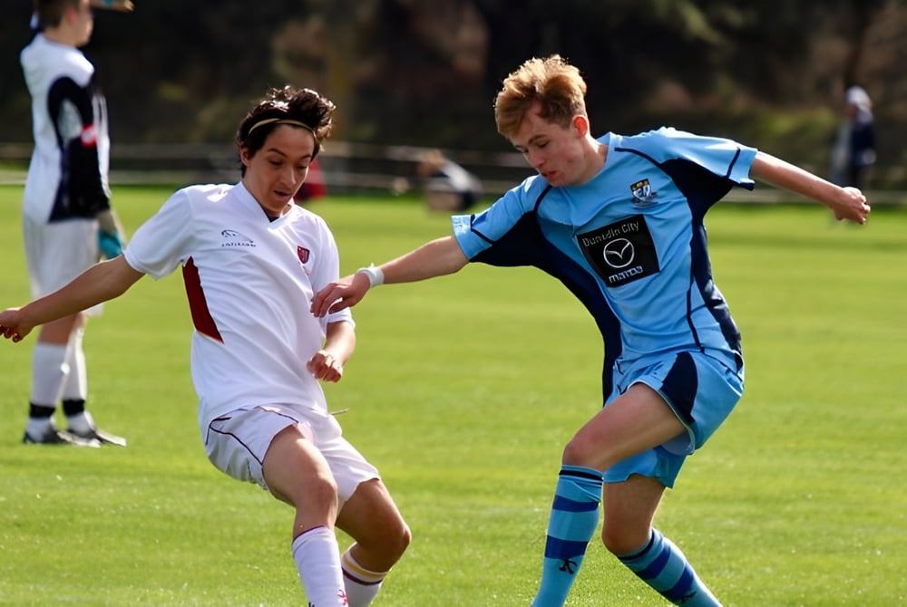 Zwei Schülerinnen und Schüler der King's High School spielen Fußball auf einem Spielfeld mit weiteren Spielern im Hintergrund.