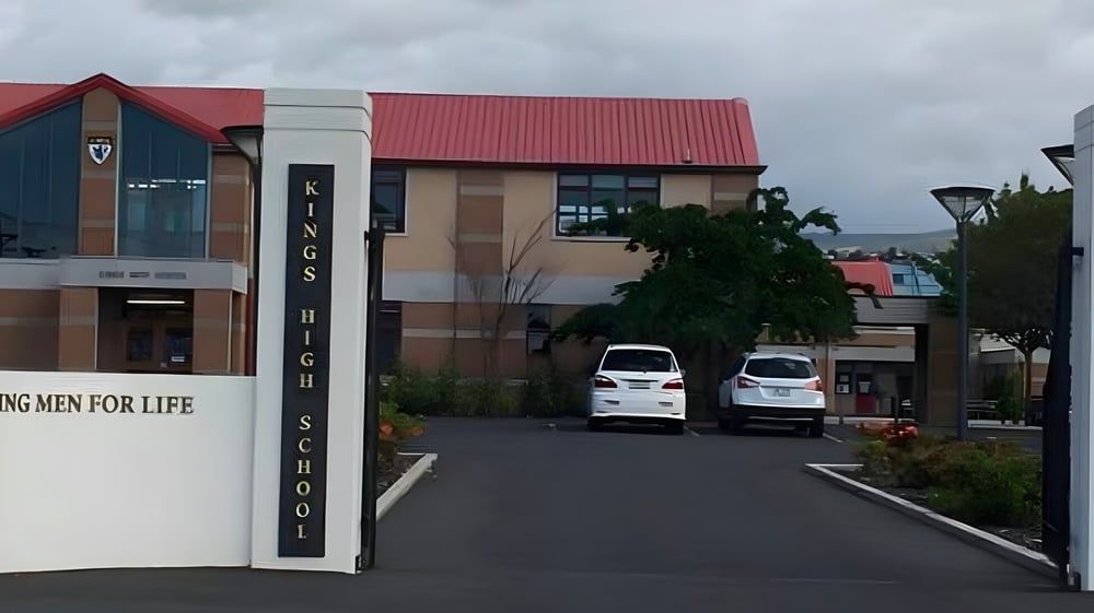 Das Gebäude der King's High School mit rotem Dach steht umgeben von Bäumen und geparkten Fahrzeugen unter bewölktem Himmel.
