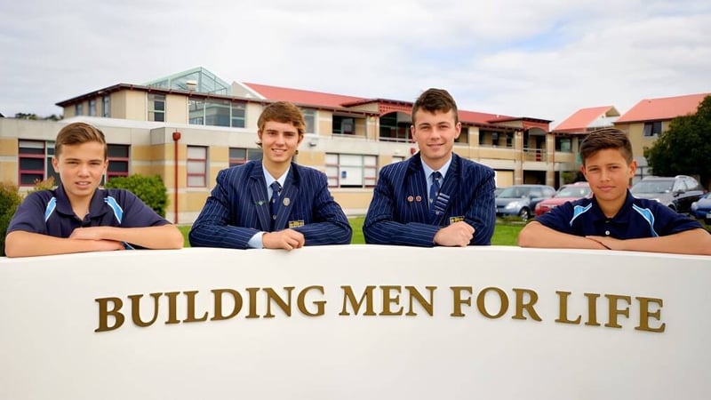 Vier Schüler der King's High School sitzen vor dem Schild 'BUILDING MEN FOR LIFE' mit Schulgebäude im Hintergrund.