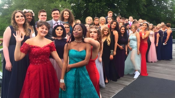 Schülerinnen der Kingston High School versammeln sich in festlicher Kleidung auf einem Holzdeck mit grüner Umgebung.