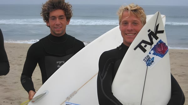 Zwei Schülerinnen der Kingston High School stehen mit Surfbrettern am Strand vor dem Meer und bewölktem Himmel.