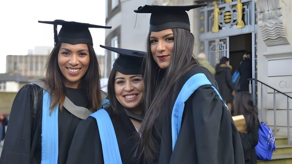 Drei Schülerinnen der Kingston Secondary School in Talaren lächeln und posieren vor einem Gebäude bei der Abschlussfeier.