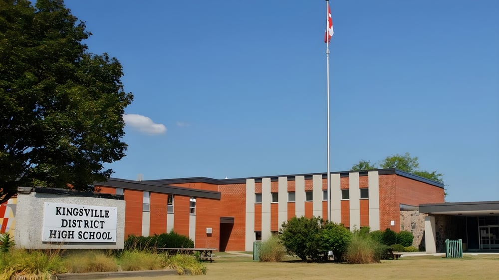 Das Backsteingebäude der Kingsville District High School steht umgeben von Bäumen und Grünflächen mit einem hohen Flaggenmast.