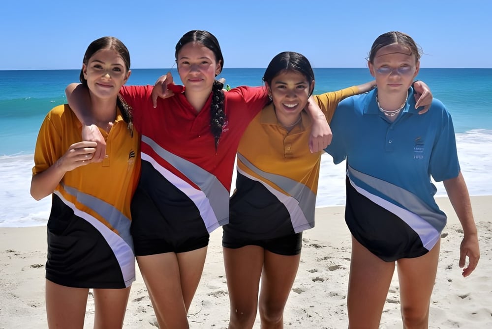 Vier Schülerinnen des Kinross College stehen gemeinsam am Sandstrand mit klarem blauem Meer im Hintergrund.