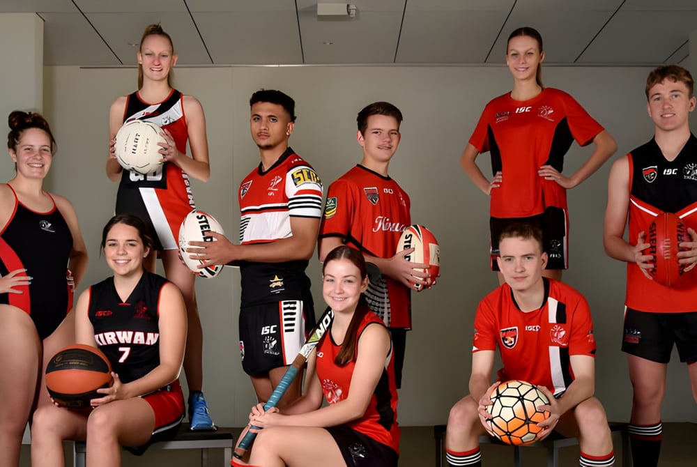 Eine Gruppe junger Athletinnen und Athleten der Kirwan State High School posiert in der Sporthalle in Teamuniformen mit Sportgeräten.