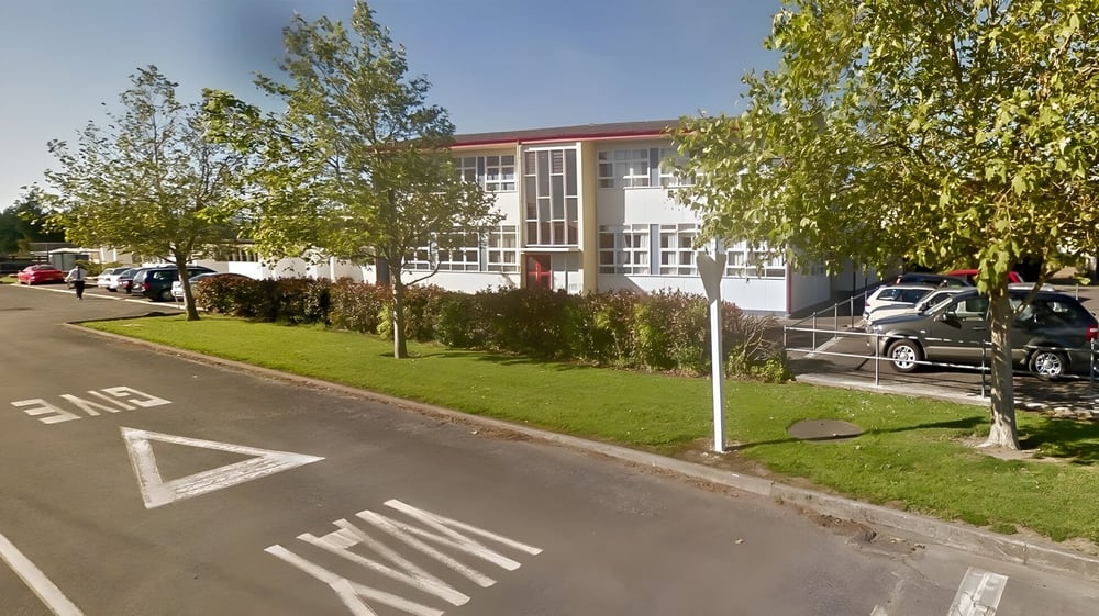 Das moderne mehrstöckige Gebäude des Kuranui College ist von Bäumen umgeben und liegt an einer Straße mit Fahrbahnmarkierungen.