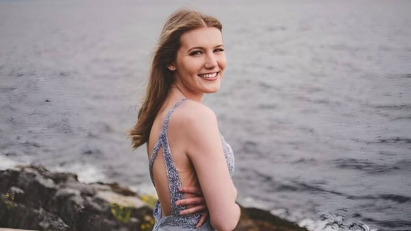 Eine junge Frau steht lächelnd vor einer felsigen Landschaft mit bewölktem Himmel auf dem Gelände der Kwalikum Secondary School.