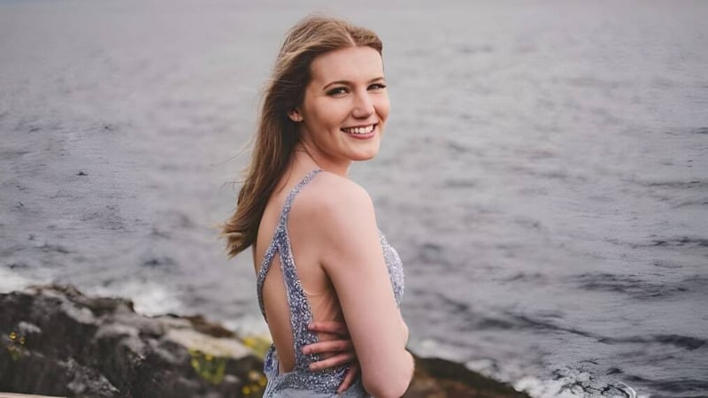 Eine junge Frau steht lächelnd vor einer felsigen Landschaft mit bewölktem Himmel auf dem Gelände der Kwalikum Secondary School.