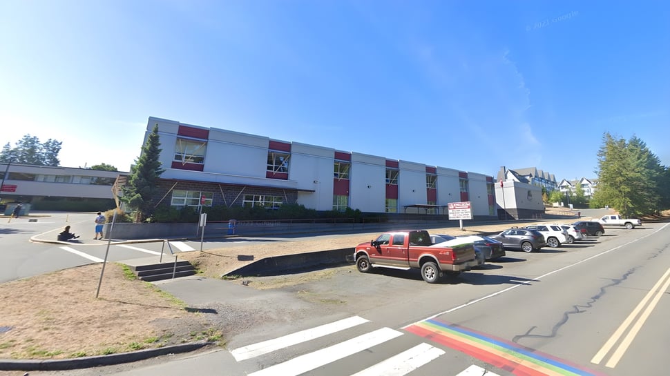 Das mehrstöckige Gebäude der Kwalikum Secondary School mit rot-weißer Fassade ist von geparkten Fahrzeugen umgeben.