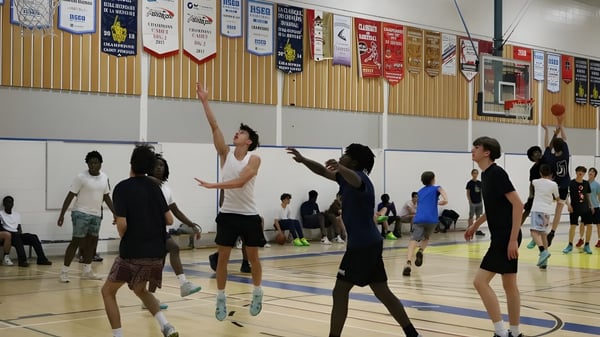 Schüler der École secondaire publique L'Académie de la Seigneurie spielen Basketball auf dem Sportplatz mit Bannern im Hintergrund.