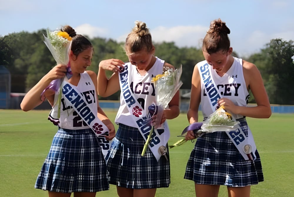 Drei Schülerinnen der Lacey Township High School in Cheerleader-Uniformen mit Pompons stehen auf einem Feld mit Bäumen und blauem Himmel.
