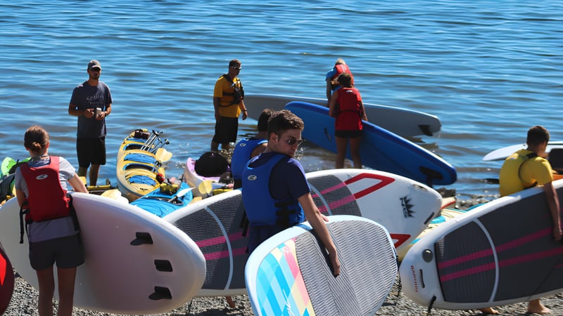 Schüler der Ladysmith Secondary School stehen auf Paddleboards auf einem Gewässer mit blauem Hintergrund.