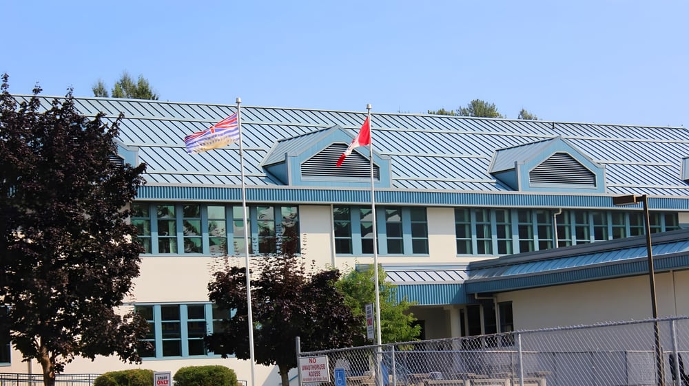 Das mehrstöckige Gebäude mit grünem Metall-Dach der Lake Cowichan Secondary School ist von Bäumen und einem Zaun umgeben.