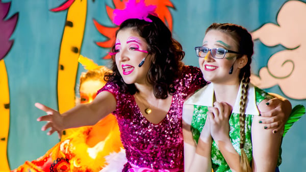 Zwei Frauen in bunten, glitzernden Outfits vor einem farbenfrohen, abstrakten Hintergrund auf dem Campus der Lake Mary Preparatory School.