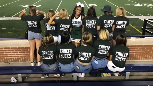 Eine Gruppe junger Frauen mit schwarzen Shirts und dem Schriftzug Cancer Sucks steht auf dem Sportfeld der Lake Orion High School.