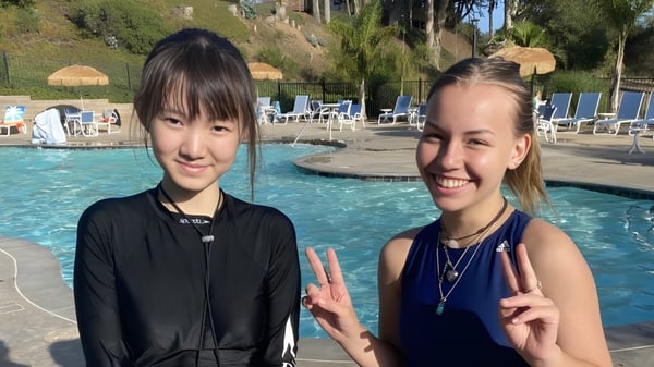 Zwei Schülerinnen der Lake Tahoe Preparatory School zeigen lächelnd Friedenszeichen vor einem Schwimmbecken mit Palmen.