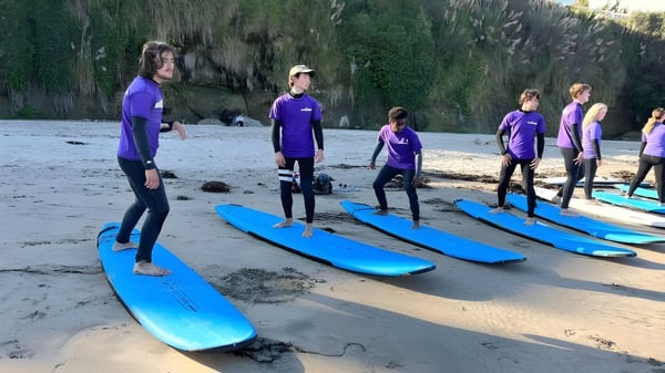 Schüler der Lake Tahoe Preparatory School stehen in lila Shirts und Neoprenanzügen am Strand neben blauen Surfboards vor einem grünen Wald.