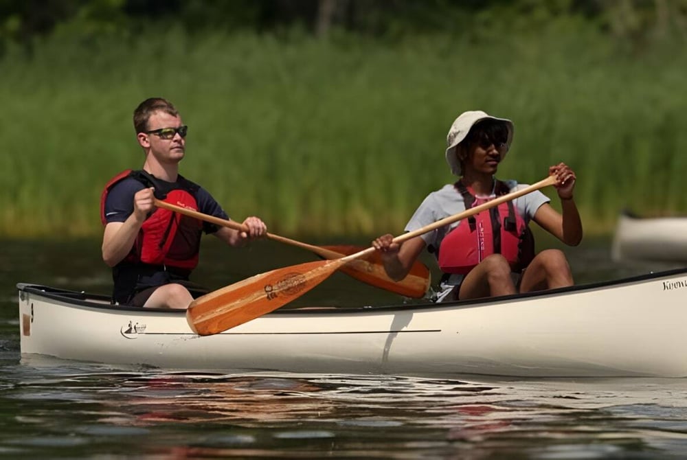 Zwei Schüler der Lakefield College School paddeln in einem Kanu auf einem ruhigen Gewässer umgeben von grüner Vegetation.