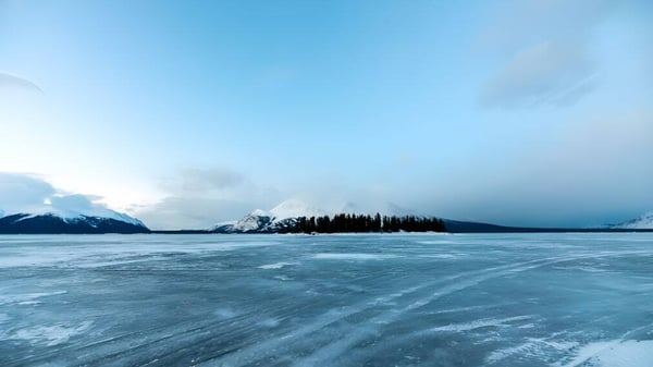 Ein zugefrorener See mit schneebedeckten Bergen und Kiefern unter bewölktem Himmel nahe der Lakes District Secondary School.