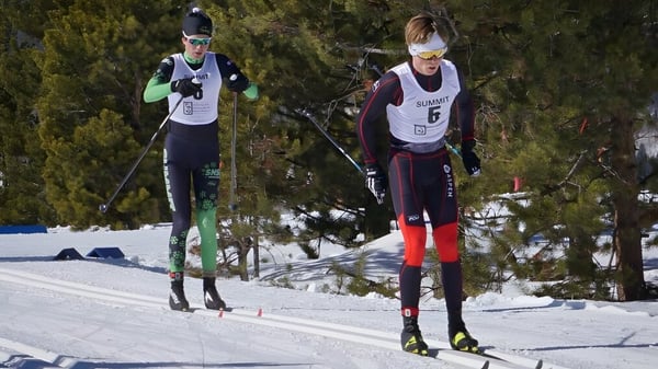 Zwei Schüler der Lakes District Secondary School sind auf einer verschneiten Loipe beim Skifahren umgeben von Kiefern.