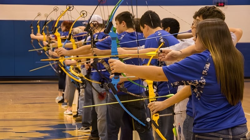 Schüler der Lakes District Secondary School stehen in blauen Uniformen in der Turnhalle und schießen mit Pfeil und Bogen auf Zielscheiben.