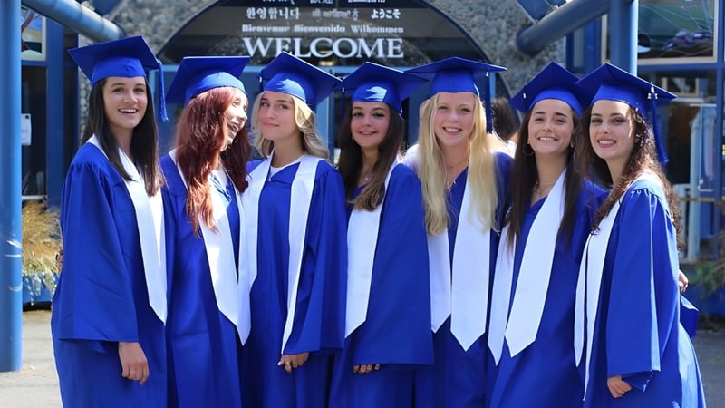 Eine Gruppe von Schülerinnen der Lambrick Park Secondary School feiert vor einem Welcome-Schild ihren Schulabschluss in blauen Talaren.