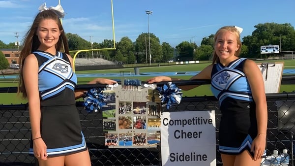 Zwei Cheerleader in blauen und weißen Uniformen stehen an der Seitenlinie der Lansing Catholic High School vor einem Sportschild.