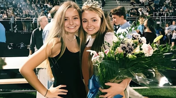 Zwei Schülerinnen der Lansing Catholic High School halten zusammen einen Blumenstrauß und posieren mit einer Menschenmenge auf einer Bühne im Hintergrund.