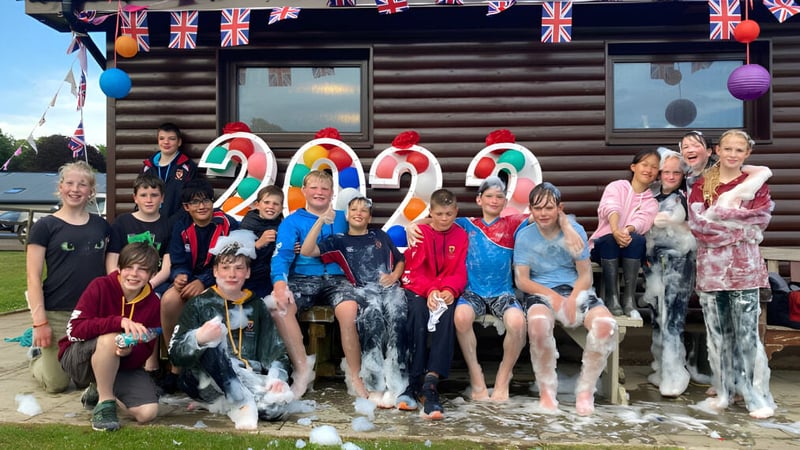 Eine Gruppe Schülerinnen und Schüler der Lathallan School versammelt sich vor einer Holzhütte mit Union Jack Fahnen nach einer bunten Festaktivität.
