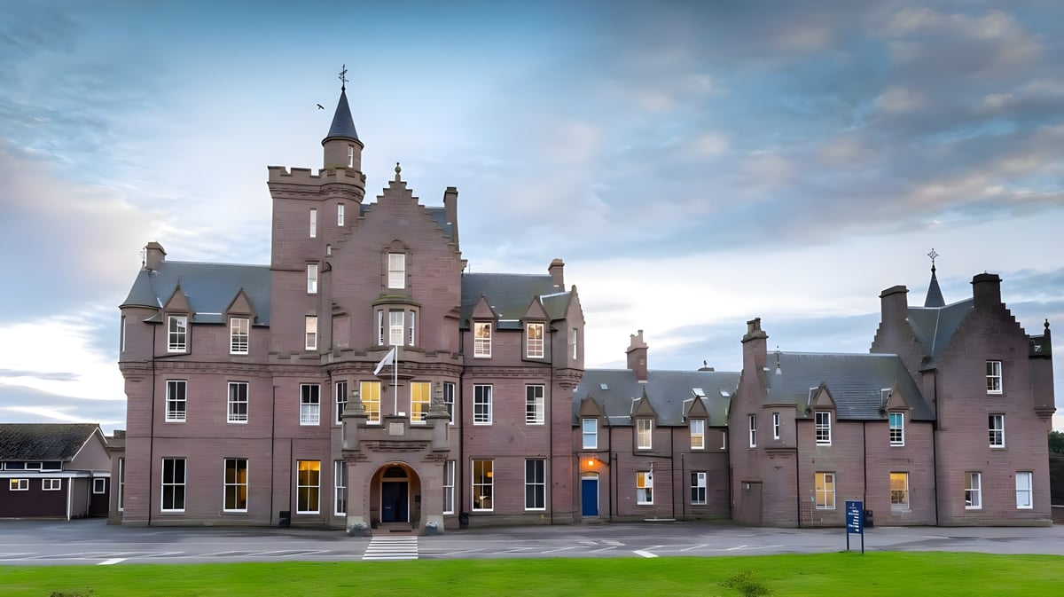 Das prächtige schlossähnliche Gebäude der Lathallan School mit Türmen steht vor kleineren Gebäuden und einer Wiese unter bewölktem Himmel.
