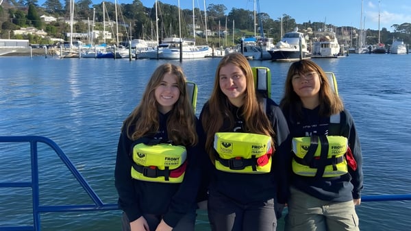 Drei Schülerinnen der Le Fevre High School tragen gelbe Schwimmwesten vor einem Hafen mit Booten.