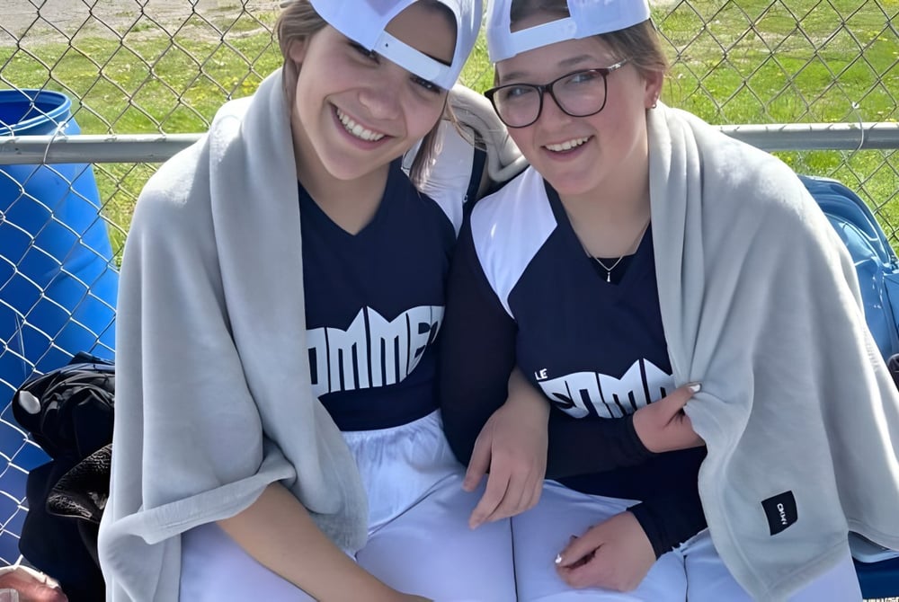 Zwei lächelnde Schülerinnen der École secondaire publique Le Sommet stehen vor einem Zaun mit blauem Container im Hintergrund.