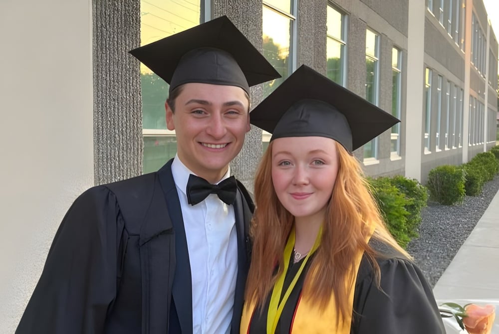 Zwei Absolventen stehen vor einem Gebäude der Leamington District Secondary School in Abschlussgewändern und Mortarboards.