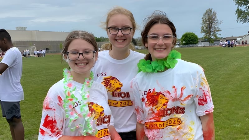 Drei Schülerinnen der Leamington District Secondary School posieren lächelnd im Freien auf einer Wiese.