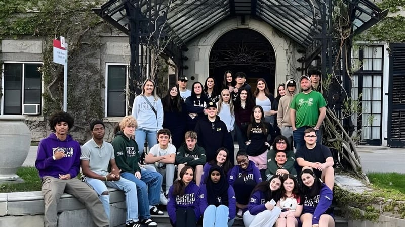 Eine Gruppe von Schülern vor dem verzierten Eingang der École secondaire publique L'Héritage mit grüner Umgebung.