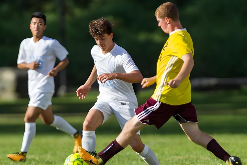 Drei Schüler der Léman Manhattan Preparatory School spielen Fußball auf einem grasbewachsenen Spielfeld.