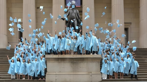 Schüler der Léman Manhattan Preparatory School versammeln sich in blauen Roben um eine große Statue auf einer Treppe.