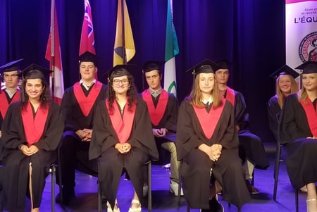 Absolventinnen und Absolventen der École élémentaire et secondaire publique L'Équinoxe stehen in akademischer Kleidung auf einer Bühne bei der Abschlussfeier.