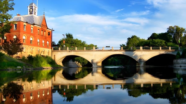 Das historische Gebäude der École élémentaire et secondaire publique L'Équinoxe mit Uhrenturm steht auf einer Brücke über einem ruhigen Fluss.