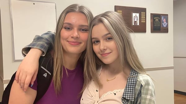 Zwei Schülerinnen mit langen blonden Haaren posieren lächelnd in einem Raum mit gerahmten Kunstwerken auf dem Campus der Lewisporte Collegiate.