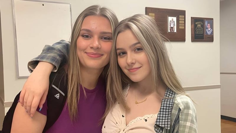 Zwei Schülerinnen mit langen blonden Haaren posieren lächelnd in einem Raum mit gerahmten Kunstwerken auf dem Campus der Lewisporte Collegiate.