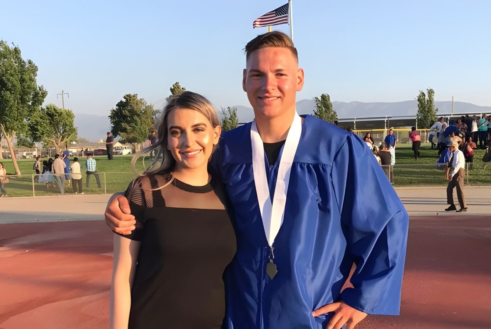 Zwei Absolventen der Littlerock High School stehen in ihren Abschlussroben und halten ihre Diplome vor einer Flagge und Bäumen.