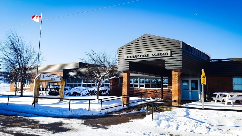 Das hölzerne Schulgebäude der Livingstone School steht in einer verschneiten Landschaft mit kahlen Bäumen und blauem Himmel.