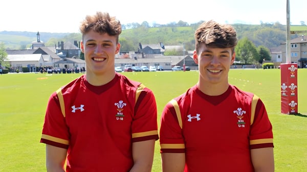 Zwei Schüler des Llandovery College stehen in roten Sporttrikots mit walisischem Flaggenemblem auf einem Rasenfeld mit Blick auf eine Stadt im Hintergrund.